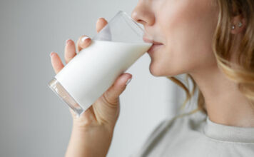 Das Close-Up einer Frau, welche ein Glas Milch trinkt. Die Milch in dem Glas ist halbvoll und die Fingernägel der Frau haben den selben milchweißen Farbton wie die Milch.
