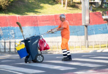 Ein älterer Mann mit grau-weißen Haaren ist in orangefarbene Arbeitsklamotten mit schwarz-weißen großen Stiefeln gekleidet und schiebt eine schwarze Mülltonne. Er arbeitet als Müllmann. An der Abfalltonne hängen Tüten und ein Eimer sowie ein Besen.