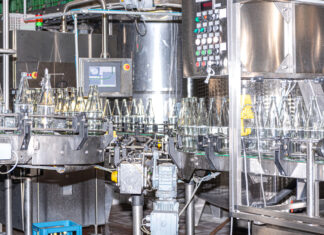 Silberne, große Maschinen im Inneren einer Fabrik waschen Glasflaschen in einer industriellen Spülmaschine. Die Flaschen stehen auf dem Fließband nah beieinander.