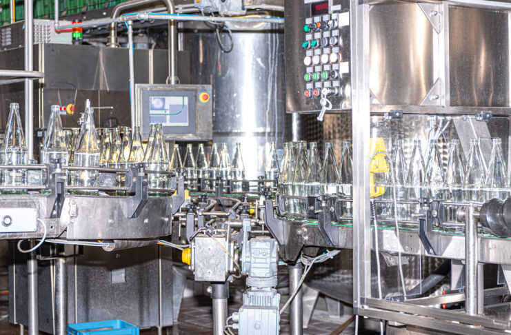 Silberne, große Maschinen im Inneren einer Fabrik waschen Glasflaschen in einer industriellen Spülmaschine. Die Flaschen stehen auf dem Fließband nah beieinander.