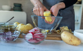 Eine Person schält eine Kartoffel über einer Schale aus Glas. Daneben stehen Zwiebeln, Gurken, Radieschen und rohe Kartoffeln. Die Person bereitet einen Kartoffelsalat vor.