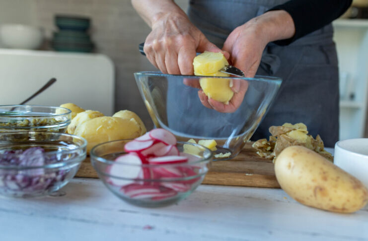 Eine Person schält eine Kartoffel über einer Schale aus Glas. Daneben stehen Zwiebeln, Gurken, Radieschen und rohe Kartoffeln. Die Person bereitet einen Kartoffelsalat vor.