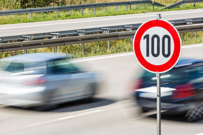 Zwei Autos fahren auf der Autobahn sehr schnell, weshalb sie unscharf und verwischt sind. Sie rasen an einem Schild vorbei, das die Geschwindigkeitsbegrenzung anzeigt. Das Tempolimit beträgt hier 100 km/h.