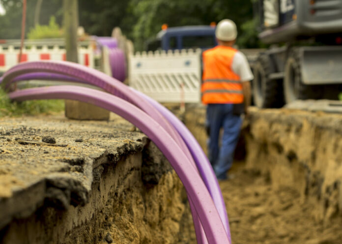 Einige Kabel ragen aus einer Baustelle und deren Grube heraus. Sie sind pink gefärbt, während im Hintergrund ein Bauarbeiter mit Helm vor einer Bauabsperrung steht.