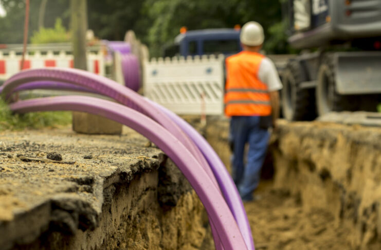 Einige Kabel ragen aus einer Baustelle und deren Grube heraus. Sie sind pink gefärbt, während im Hintergrund ein Bauarbeiter mit Helm vor einer Bauabsperrung steht.