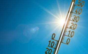 Ein Thermometer wird in die Sonne gehalten. Der Hintergrund ist ein blauer Himmel, ohne Wolken. Die Sonne ist ebenfalls hinter dem Thermometer zu sehen.