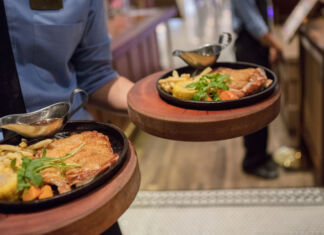 Ein Keller in Uniform in einem Restaurant bringt zwei Teller an eine Tisch. Auf den Tellern befinden sich Schnitzel, Pommes und Gemüse.