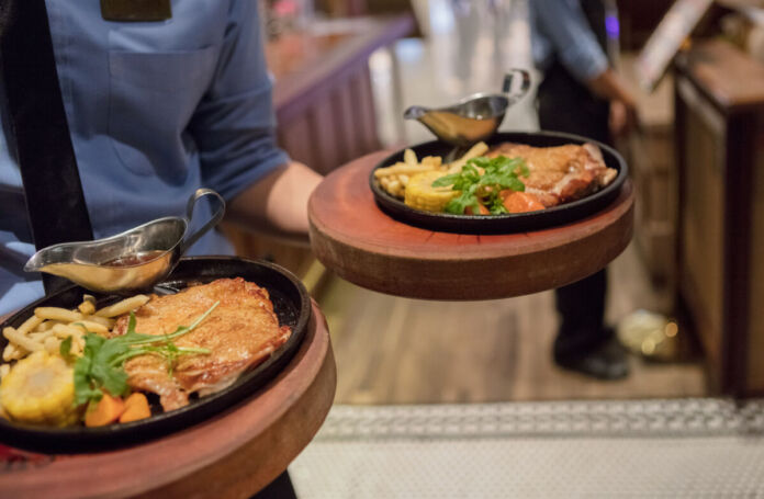 Ein Kellner in Uniform in einem Restaurant bringt zwei Teller an einen Tisch. Auf den Tellern befinden sich Schnitzel, Pommes und Gemüse.