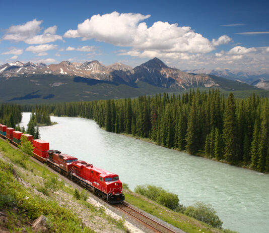 Ein Güterzug fährt mit vielen Waggons durch eine malerische Landschaft an einem Fluss und Wald entlang bis hin zum Meer.