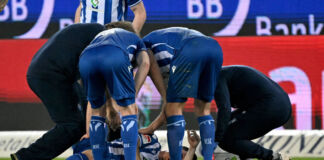 Ein Spieler des Karlsruher SC liegt verletzt am Boden. Er krümmt sich vor Schmerzen. Seine Mannschaftskameraden beugen sich zu ihm herunter und halten ihn mit den Armen fest. Sie versuchen ihm zu helfen. Die Spieler befinden sich noch auf dem Spielfeld.