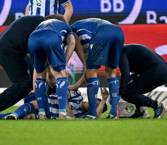 Ein Spieler des Karlsruher SC liegt verletzt am Boden. Er krümmt sich vor Schmerzen. Seine Mannschaftskameraden beugen sich zu ihm herunter und halten ihn mit den Armen fest. Sie versuchen ihm zu helfen. Die Spieler befinden sich noch auf dem Spielfeld.