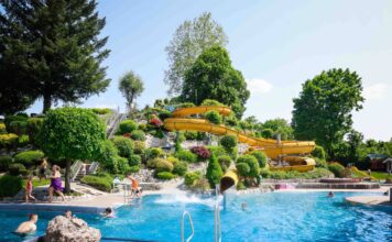 Ein großes Spaßbad oder ein Freibad im Sommer bei schönem Wetter. Im Hintergrund befindet sich eine große gelbe Wasserrutsche mit mehreren Drehungen für große und kleine Besucher.