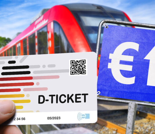 Eine Hand hält ein Deutschland-Ticket. Daneben befindet sich ein blaues Schild mit einem Euro-Zeichen und einem Pfeil darauf. Im Hintergrund fährt ein Zug.