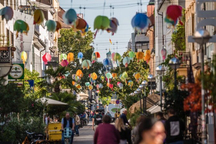 Mehrere Bürger und Passanten gehen durch eine bunt geschmückte Straße in Karlsruhe an einem schönen Tag. Es findet gerade ein großes Straßenfest oder ein Kulturfest statt, weshalb so viele Menschen unterwegs sind.