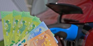 Im Vordergrund, vor einer modernen Ladesäule für E-Autos, hält jemand eine beträchtliche Anzahl von Geldscheinen in seiner Hand. Das Laden von Elektroautos an Ladestationen wird immer teurer.
