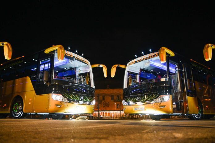 Zwei Busse stehen nebeneinander in der Nacht, beide Reisebusse haben das Licht angeschaltet.
