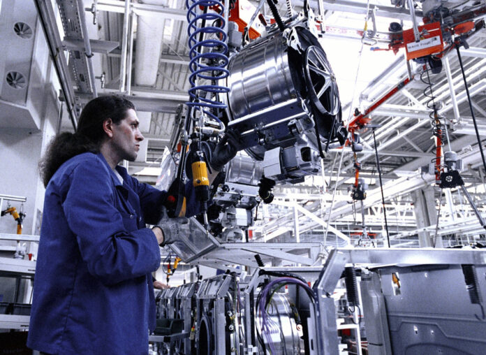 Ein Mitarbeiter in einem Blaumann bedient in einer großen Fabrikhalle eine schwere Maschine. Es scheint eine Art Motor zu sein, der an einem Kran hängt und den der Arbeiter vorsichtig manövriert.