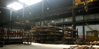 In einer großen Lagerhalle befinden sich viele Stahlrohre unterschiedlicher Länge und mit unterschiedlichem Durchmesser in großen Regalen. Über den Rohren bewegt sich eine Maschine.