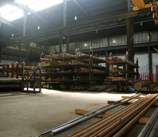 In einer großen Lagerhalle befinden sich viele Stahlrohre unterschiedlicher Länge und mit unterschiedlichem Durchmesser in großen Regalen. Über den Rohren bewegt sich eine Maschine.