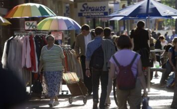 Viele Passanten schlendern durch die Sonne, einige halten einen bunten Sonnenschirm in der Hand. Sie kaufen in einigen Geschäften der Innenstadt ein und betrachten die Auslagen der Händler.