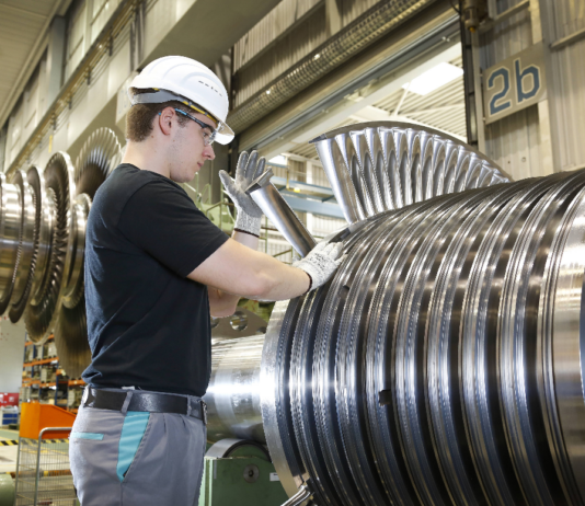 Ein Arbeiter steht vor einer riesigen Anlage in einer großen Industriehalle und legt seine Hand auf etwas, das aus Stahlringen besteht.
