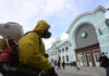 Ein Mann in einem kompletten Schutzanzug mit einer Gasmaske desinfiziert mit einem Gerät eine Bahnhofsstation. Im Hintergrund laufen einige Leute in und aus dem Bahnhof.