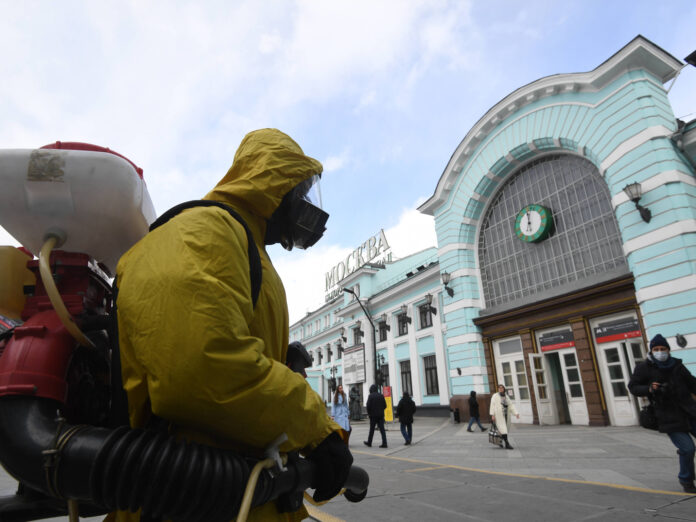 Ein Mann in einem kompletten Schutzanzug mit einer Gasmaske desinfiziert mit einem Gerät eine Bahnhofsstation. Im Hintergrund laufen einige Leute in und aus dem Bahnhof.