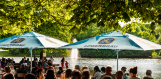 Ein Biergarten in Baden-Württemberg. Im Schatten unter den großen Sonnenschirmen sitzen sehr viele Gäste. Sie nehmen Speisen ein oder trinken Bier und andere Getränke.