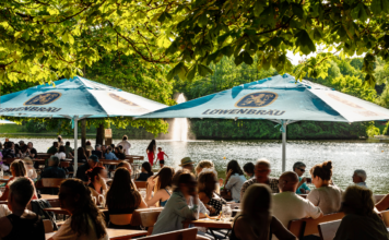 Ein Biergarten in Baden-Württemberg. Im Schatten unter den großen Sonnenschirmen sitzen sehr viele Gäste. Sie nehmen Speisen ein oder trinken Bier und andere Getränke.
