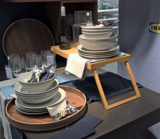 Gestapelte weiße, schicke Teller, Gabel, Messer und Löffel von IKEA liegen auf mehreren Holztischen und sind für Kunden des Möbel-Einrichtungshauses zum Kauf verfügbar.