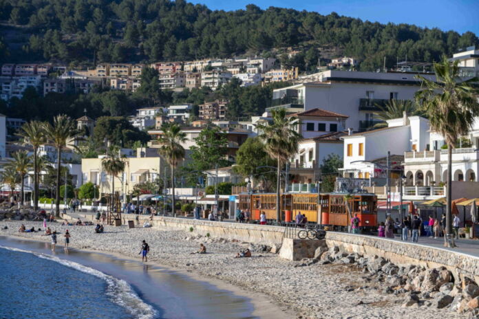 Ein Strand an einer Küste, die von zahlreichen Häusern im Hintergrund und auf Waldhügeln bebaut wurde. Wellen brechen am Sandstrand und Touristen gehen vereinzelt ins Wasser um sich abzukühlen.