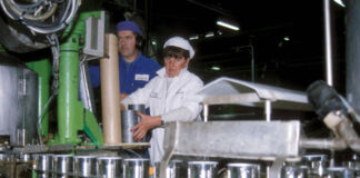 Ein Blick in eine Fabrik. Eine Frau und ein Mann stehen als Angestellte an einem einem Fließband und stellen Konservendosen her.
