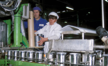 Ein Blick in eine Fabrik. Eine Frau und ein Mann stehen als Angestellte an einem einem Fließband und stellen Konservendosen her.