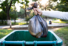 Eine Person wirft einen Müllsack in eine grüne Mülltonne. Es handelt sich vermutlich um Hausmüll oder gemischten Müll. Der Müllsack ist schwarz und teilweise durchsichtig.
