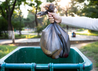 Eine Person wirft einen Müllsack in eine grüne Mülltonne. Es handelt sich vermutlich um Hausmüll oder gemischten Müll. Der Müllsack ist schwarz und teilweise durchsichtig.