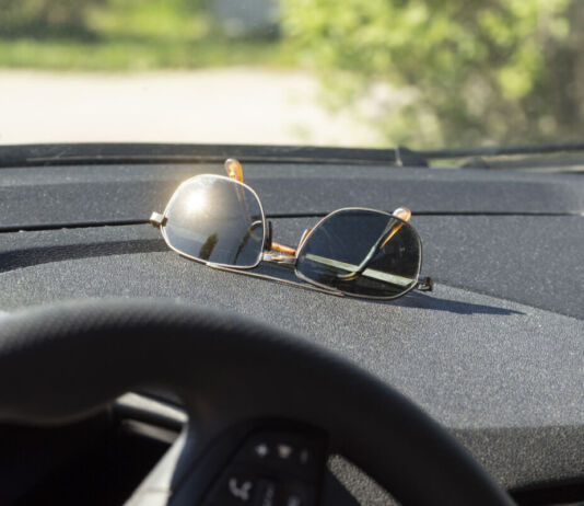 Eine Sonnenbrille liegt unter der Windschutzscheibe im Auto im Cockpit. Die Sonne scheint darauf und ein Glas blendet die Sonne drastisch zurück.