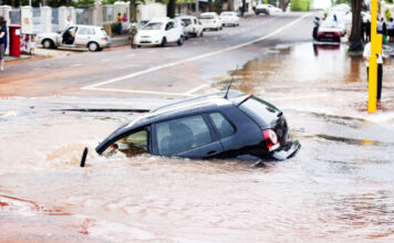 Ein Auto treibt in einer Sturzflut. Die Straße ist völlig überschwemmt, sodass das Wasser braun und dreckig ist. Es regnet und auch weitere Teile der Straße stehen bereits unter Wasser.
