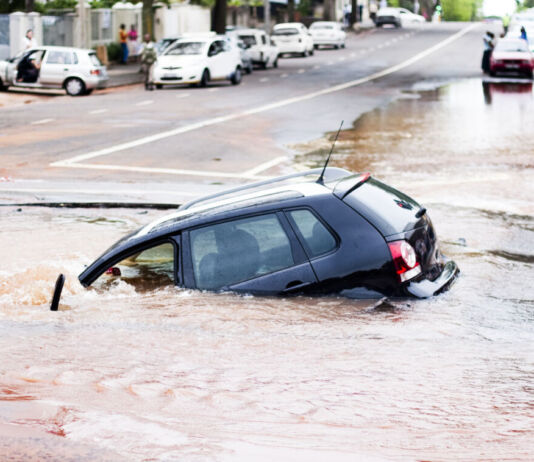 Ein Auto treibt in einer Sturzflut. Die Straße ist völlig überschwemmt, sodass das Wasser braun und dreckig ist. Es regnet und auch weitere Teile der Straße stehen bereits unter Wasser.