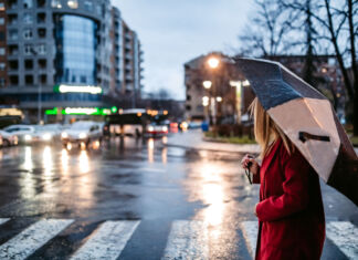 Eine Frau mit einem Regenschirm überquert eine Straße. Im Hintergrund sieht man einige Autos. Es dämmert bereits und regnet leicht.