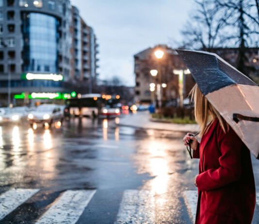 Eine Frau mit einem Regenschirm überquert eine Straße. Im Hintergrund sieht man einige Autos. Es dämmert bereits und regnet leicht.
