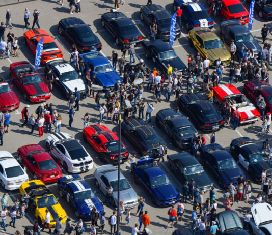 Auf einem riesig großen Parkplatz stehen viele Ford Mustangs unterschiedlicher Jahre. Eine Menschenmasse läuft zwischen den einzelnen Modellen entlang und schaut sich jedes an.