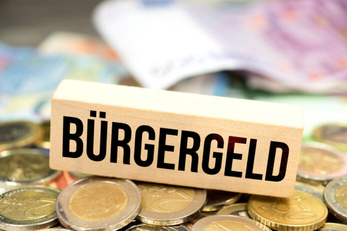 Bargeld, vor allem zwei Euro Münzen und verschiedene Geldscheine, liegen auf einem Tisch. Darauf liegt ein Schild aus Holz mit der Aufschrift Bürgergeld