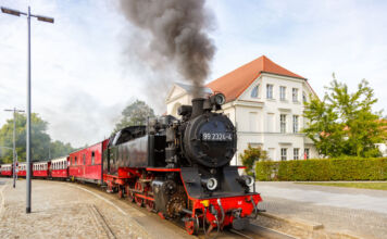 Eine historische schwarze Dampflokomotive fährt mit schwarzem Dampf an einem Bahnhof vorbei, das Wetter ist sonnig und lädt zu Ausflügen mit der Lokomotive ein. Alte Eisenbahnen haben viele Fans.