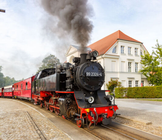 Eine historische schwarze Dampflokomotive fährt mit schwarzem Dampf an einem Bahnhof vorbei, das Wetter ist sonnig und lädt zu Ausflügen mit der Lokomotive ein. Alte Eisenbahnen haben viele Fans.