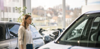 Eine erwachsene Frau steht in dem Gebäude eines Autohändlers und schaut sich nach einem Neuwagen um. Sie steht vor einem großen SUV. Im Hintergrund erkennt man aus den großen Fenstern heraus eine kleine Stadt.