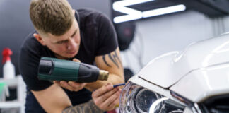 Ein Mitarbeiter einer Werkstatt klebt etwas mit einem Heißkleber und Werkzeug an einem Scheinwerfer eines Autos fest. Das Fahrzeug ist weiß und der Mitarbeiter konzentriert sich auf die Arbeit.