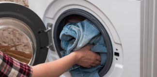 Eine Frau legt ihre Wäsche in eine weiße Waschmaschine. Sie trägt ein Flanell-Hemd und wirft Jeans in die Maschine, um sie zu waschen.