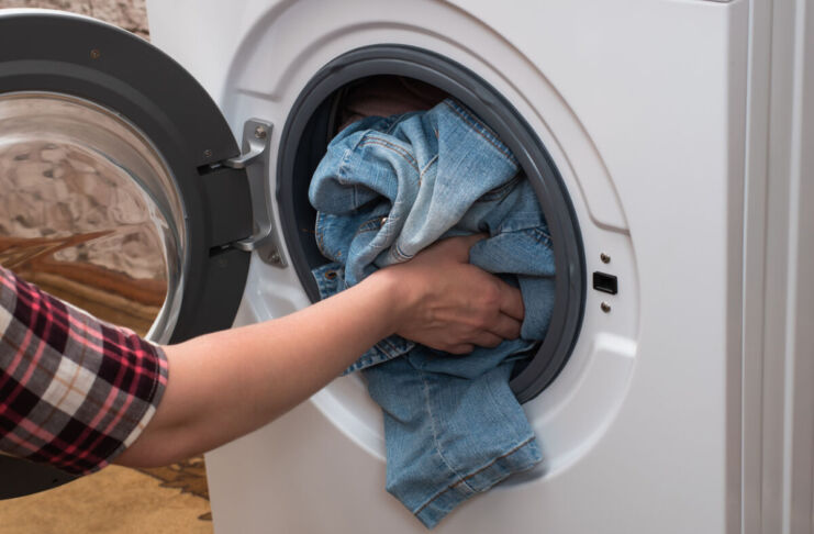 Eine Frau legt ihre Wäsche in eine weiße Waschmaschine. Sie trägt ein Flanell-Hemd und wirft Jeans in die Maschine, um sie zu waschen.