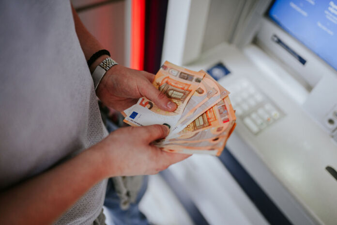 Junge Männerhände halten mehrere 50-Euro-Scheine fest. Der Mann steht vor einem Geldautomaten und zählt sein Geld, welches er gerade abgehoben hat, nach. An seinem linken Handgelenk befindet sich eine Uhr.