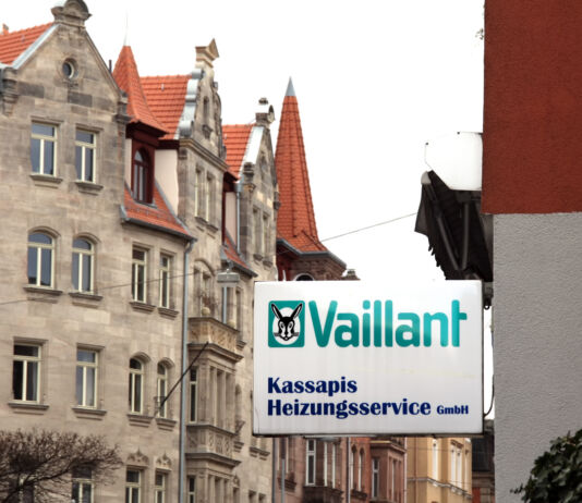 Eine Straße mit historisch anmutenden Gebäuden unter bedecktem Himmel. An einem der Hauswände hängt das Schild der Firma Vaillant in den klassischen Logofarben des Unternehmens.
